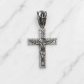 A silver-toned crucifix pendant with a detailed corpus and halo, set with small clear stones, displayed on a white marble background.