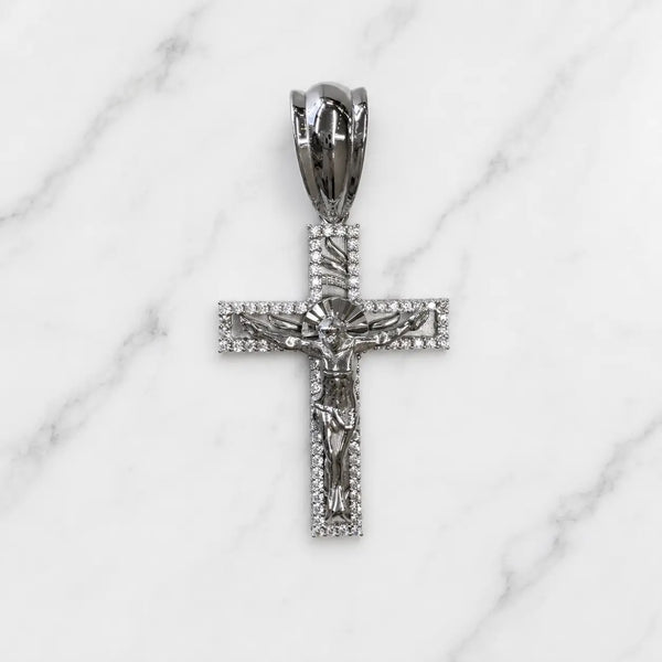 A silver-toned crucifix pendant with a detailed corpus and halo, set with small clear stones, displayed on a white marble background.