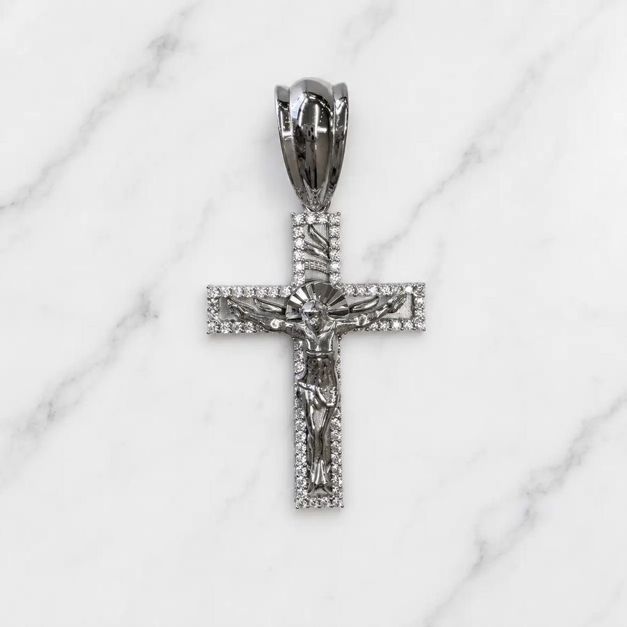 A silver-toned crucifix pendant with a detailed corpus and halo, set with small clear stones, displayed on a white marble background.