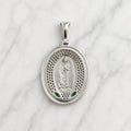 A silver oval pendant featuring a relief of Our Lady of Guadalupe, surrounded by a perforated border with small green and red accents, displayed on a white marble background.