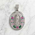 An oval silver pendant featuring a central silver figure, surrounded by pink and green gemstone accents, displayed on a white marble background.