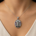 A close-up of a silver oval pendant necklace worn on a model's neck. The pendant features a detailed silver figure in the center, surrounded by a border of small clear stones and accents of pink and green gemstones.