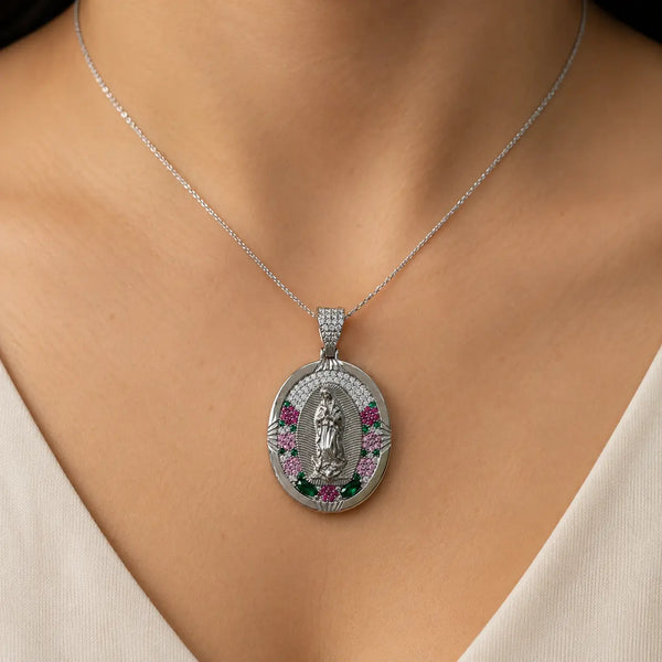 A close-up of a silver oval pendant necklace worn on a model's neck. The pendant features a detailed silver figure in the center, surrounded by a border of small clear stones and accents of pink and green gemstones.