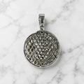 A round silver pendant with a textured, diamond-patterned openwork center and a polished bail, displayed on a white marble surface.
