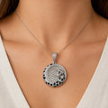 A close-up of a model's neck and chest wearing a silver-toned pendant necklace. The pendant is round and heavily embellished with clear stones, featuring a black and white spotted border with small red gemstone accents.