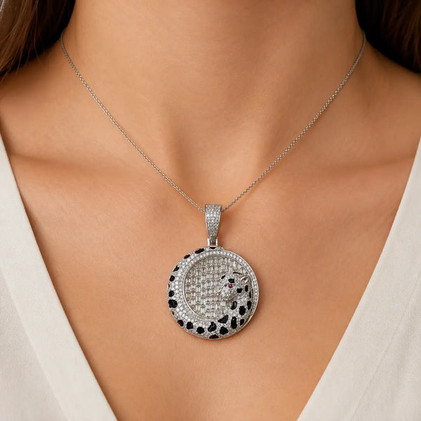 A close-up of a model's neck and chest wearing a silver-toned pendant necklace. The pendant is round and heavily embellished with clear stones, featuring a black and white spotted border with small red gemstone accents.