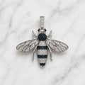 A bee-shaped pendant necklace crafted from silver-toned metal, pavé-set with clear stones on the wings and body, and accented with dark blue stones on the thorax and abdomen, displayed on a white marble background.