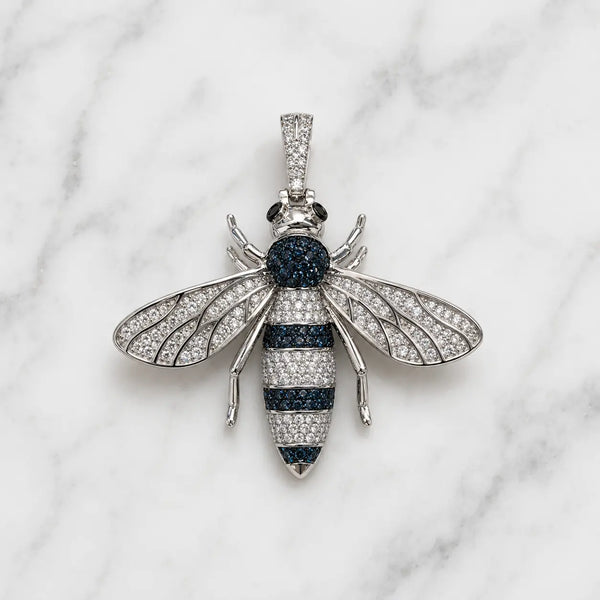 A bee-shaped pendant necklace crafted from silver-toned metal, pavé-set with clear stones on the wings and body, and accented with dark blue stones on the thorax and abdomen, displayed on a white marble background.
