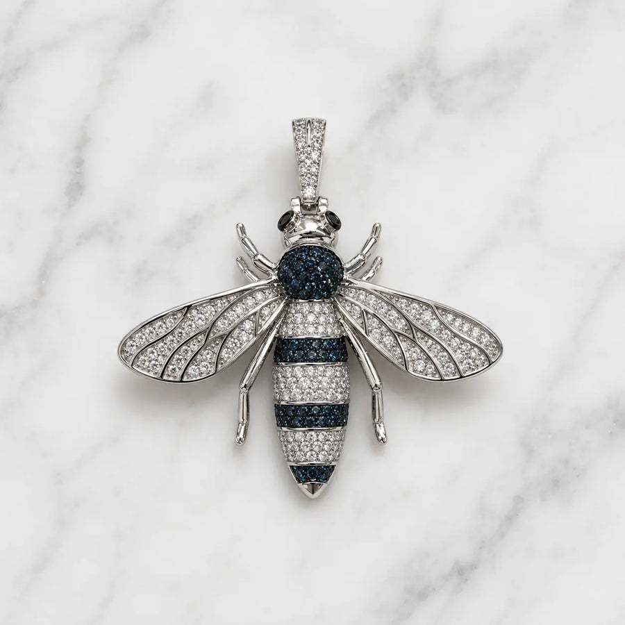 A bee-shaped pendant necklace crafted from silver-toned metal, pavé-set with clear stones on the wings and body, and accented with dark blue stones on the thorax and abdomen, displayed on a white marble background.
