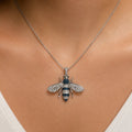 A silver-tone bee pendant necklace on a delicate chain, worn by a model. The bee pendant is detailed with clear pavé stones and features dark blue gemstone accents on its abdomen and thorax.
