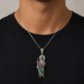 A silver-toned pendant necklace worn by a person in a black t-shirt. The pendant features a detailed figure with a silver halo and a draped sash embellished with green and pink stones.