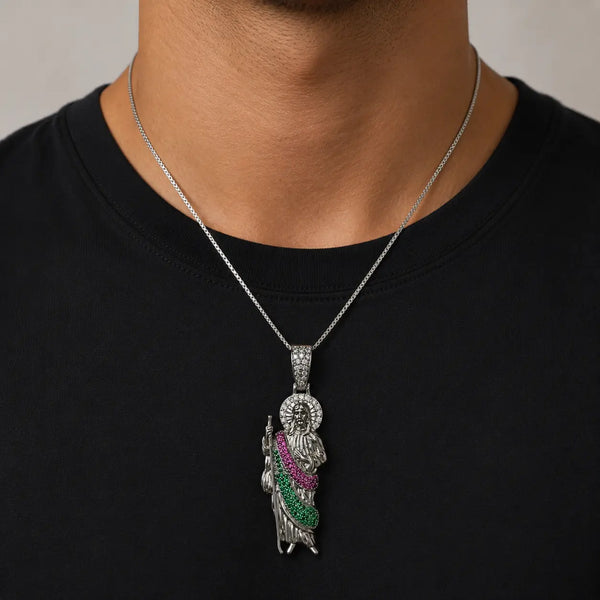 A silver-toned pendant necklace worn by a person in a black t-shirt. The pendant features a detailed figure with a silver halo and a draped sash embellished with green and pink stones.